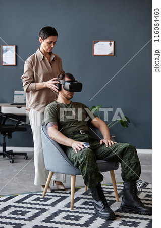 Vertical full length portrait of female psychologist setting up VR headset on army veteran during therapy session dealing with trauma and PTSD 116084463