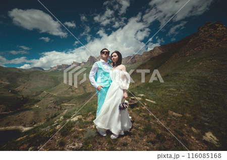 Happy bride and groom hold each other hugging while looking at the sky against the backdrop of hills and mountains. Bride in a fashionable wedding dress with artificial flowers 116085168
