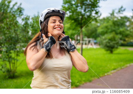 Portrait of roller skater woman putting on helmet in inline skates before rollerblading in park. Outdoor activities. Summer roller skating. 116085409