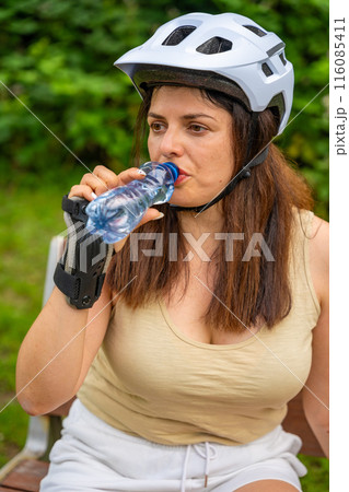 Portrait of a female roller skater drinking water while sitting on a bench in the park after roller skating. Outdoor activities. Summer roller skating. 116085411
