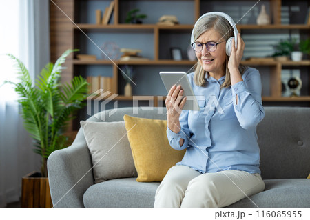 Elderly woman enjoying technology, listening to music or a podcast with headphones and using a tablet. She is sitting comfortably on a sofa in a contemporary living room setting. 116085955