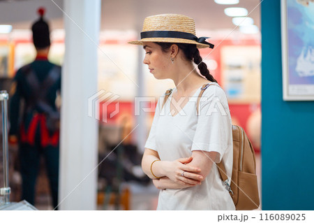 Historic education. Side view of young Caucasian woman student wearing straw hat admiring paintings in the gallery. Concept of visit museum and cultural education 116089025