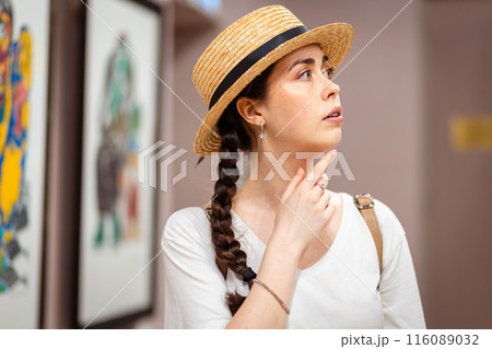 Portrait of young dreamy Caucasian woman in straw hat admiring paintings in the gallery. Concept of cultural education at museum 116089032