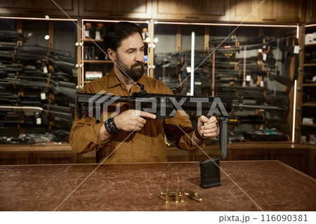 Salesman presenting new model of military gun standing at counter desk Salesman presenting new model of military gun standing at counter desk 116090381