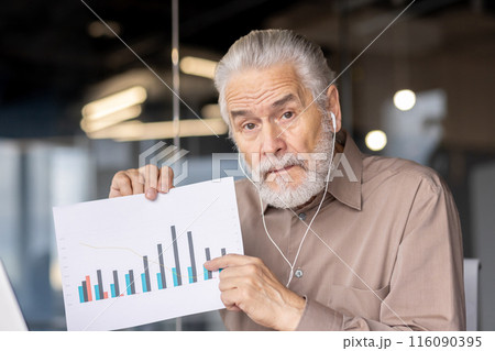 Senior businessman presenting a graph chart during a meeting in a modern office setting. The man, wearing earphones, appears to be explaining financial or business data 116090395