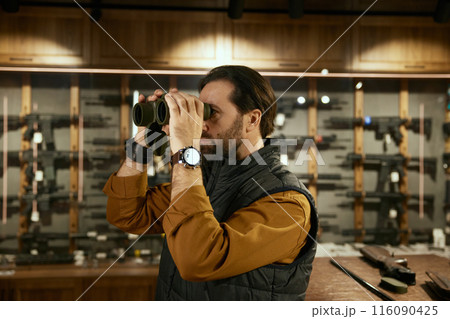 Male client choosing new binocular for hunting in weapon shop Male client choosing new binocular for hunting in weapon shop 116090425