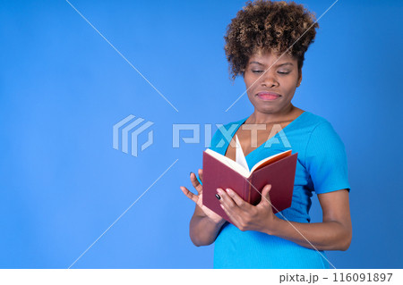 Young black woman reading a book wearing a blue dress on a blue background with copy space. Concept studying. 116091897