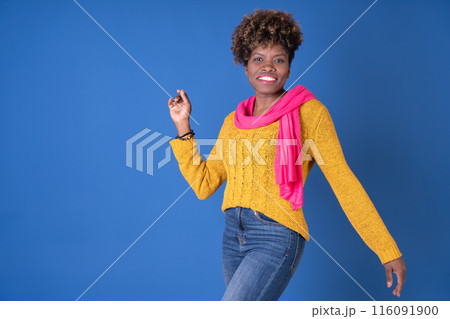 Young pretty black woman dancing with afro curly hair smiling in fuchsia scarf and yellow sweater on blue background with copy space. Happiness concept. 116091900