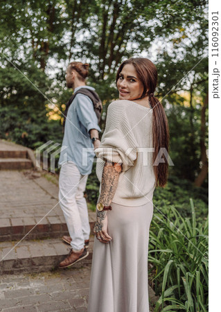 Outdoor shot of young couple in love walking on pathway through grass field. Young couple strolling in the park. Trendy couple with tattoos walking in a park. Botanic Garden. Outdoor shot of young couple in love walking on pathway through grass field. Young couple strolling in the park. Trendy couple with tattoos walking in a park. Botanic Garden. 116092301