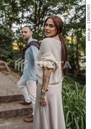 Outdoor shot of young couple in love walking on pathway through grass field. Young couple strolling in the park. Trendy couple with tattoos walking in a park. Botanic Garden. Outdoor shot of young couple in love walking on pathway through grass field. Young couple strolling in the park. Trendy couple with tattoos walking in a park. Botanic Garden. 116092303