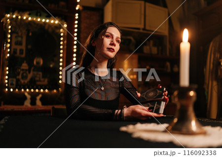 Mysterious wizard with black nails shuffling deck of tarot cards by light of burning candles. Mysterious clairvoyant performing esoteric rite, forecasting fate. Concept of esoteric, fortune telling. 116092338