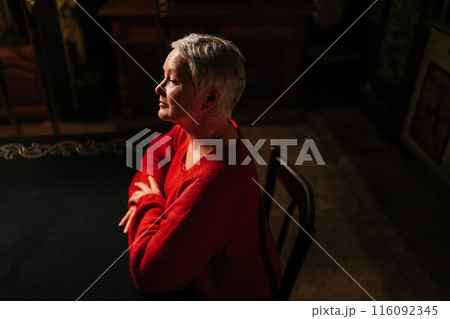 High-angle view of elegant gray-haired mature adult woman in casual red sweater sitting at table in dark room by light of burning candle, pensive looking away. High-angle view of elegant gray-haired mature adult woman in casual red sweater sitting at table in dark room by light of burning candle, pensive looking away. 116092345