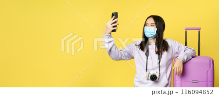 Happy tourist, asian girl on vacation taking selfie in medical mask near suitcase, photographing on smartphone, yellow background Happy tourist, asian girl on vacation taking selfie in medical mask near suitcase, photographing on smartphone, yellow background 116094852