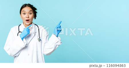Shocked asian doctor, nurse in uniform, pointing right and gasping surprised, big news, standing over blue background 116094863
