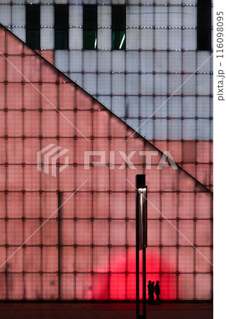 A family walking past the illuminated walls of Chongqing grand theatre in the night, an abstract yet cyberpunk scene. 116098095