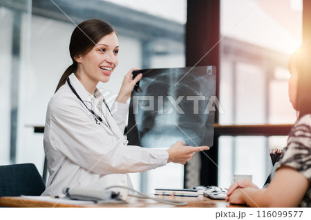 A female doctor in a white coat showing an X-ray to a patient in a modern medical office with sunlight streaming through the windows. A female doctor in a white coat showing an X-ray to a patient in a modern medical office with sunlight streaming through the windows. 116099577