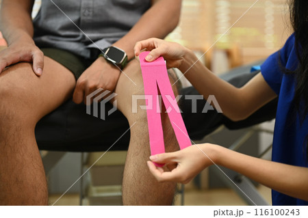 Cropped shot female physiotherapist applying pink kinesio tape on patient knee. Physical therapy and rehabilitation concept 116100243