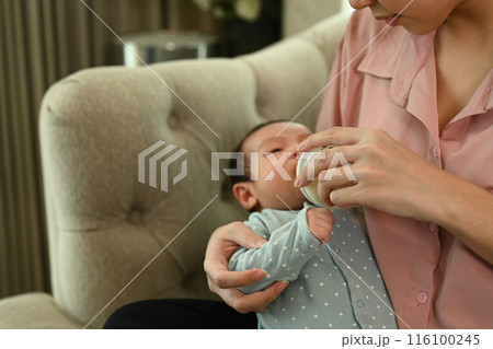 Caring mother feeding her baby with bottle and in living room on the sofa. Healthy infant nutrition concept 116100245