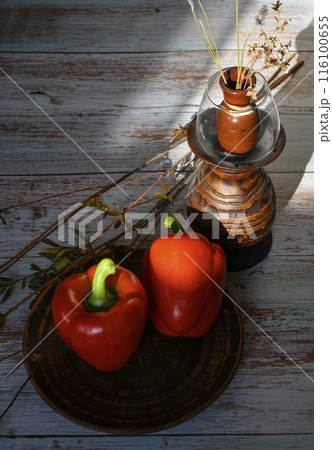 Red peppers on a clay plate, branches of wild rose and a wooden vase with dried flowers Red peppers on a clay plate, branches of wild rose and a wooden vase with dried flowers 116100655