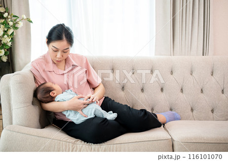 A young Asian mom comforting her newborn baby son to sleep on the couch together in the living room. A young Asian mom comforting her newborn baby son to sleep on the couch together in the living room. 116101070