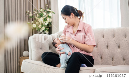 A caring Asian mom is holding her newborn baby son in her arm and feeding him milk from a bottle. 116101087