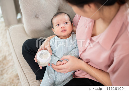 An adorable, innocence Asian newborn baby son in blue pajamas is in his mother embrace on the sofa. 116101094