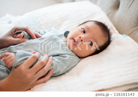 An adorable Asian newborn baby son in blue pajamas is in his bed, talking and playing with his mom. 116101143