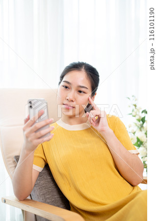 A thoughtful Asian woman is reading messages on her smartphone while relaxing on an armchair at home A thoughtful Asian woman is reading messages on her smartphone while relaxing on an armchair at home 116101190