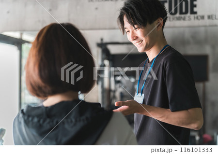 ジムで女性会員に運動メニューの説明・指導をするトレーナー・インストラクターの若いアジア人男性 116101333