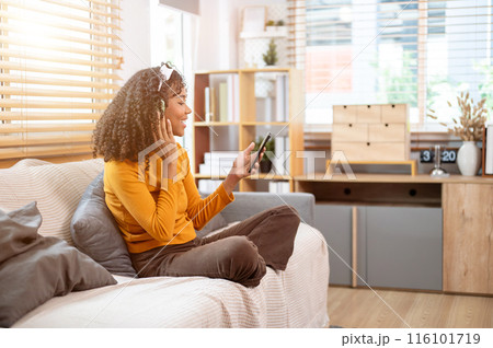 A cheerful, positive Black woman is using her phone and enjoying listening to music on the couch. 116101719