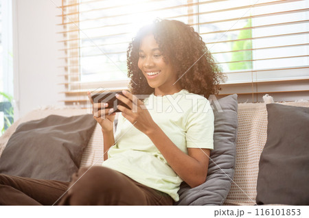 A cheerful, young Black woman is enjoying playing a mobile game on a couch in the living room. 116101853