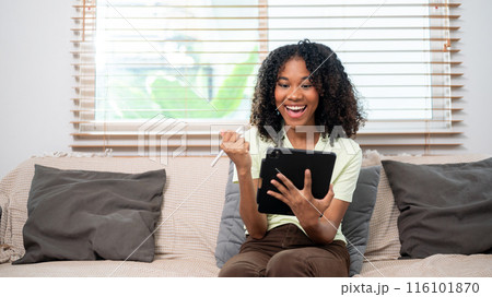 A cheerful Black woman is exciting about something on her tablet, using tablet on couch at home. 116101870