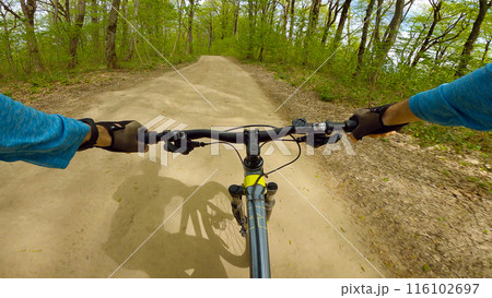 First-person view of a cyclist riding a mountain bike on a dirt trail. The cyclist is wearing a blue shirt and black gloves. The trail is surrounded by lush greenery and trees. First-person view of a cyclist riding a mountain bike on a dirt trail. The cyclist is wearing a blue shirt and black gloves. The trail is surrounded by lush greenery and trees. 116102697