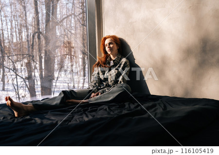 Tired pensive young woman sitting by window on sunny day on windowsill with winter nature background, feels upset, having melancholic mood, thinking about life troubles. Loneliness concept. Tired pensive young woman sitting by window on sunny day on windowsill with winter nature background, feels upset, having melancholic mood, thinking about life troubles. Loneliness concept. 116105419