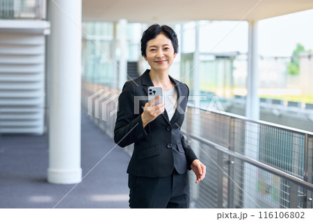 オフィスの廊下でスマホを持って立つスーツ姿のビジネスウーマン オフィスの廊下でスマホを持って立つスーツ姿のビジネスウーマン 116106802