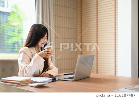 Beautiful young businesswoman using laptop and having coffee at desk in a modern office Beautiful young businesswoman using laptop and having coffee at desk in a modern office 116107497