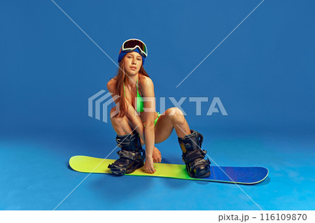 Girl combining summer swimwear and winter sports gear, ready for a unique snowboarding experience against blue studio background 116109870