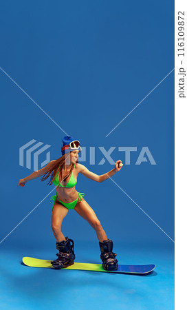Stylish woman in bikini and winter sports gear, demonstrating her adventurous spirit and love for snowboarding against blue studio background 116109872