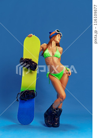 Young girl in green bikini, wearing snowboard boots and holding snowboard, blending summer style with winter sports, against blue studio background 116109877