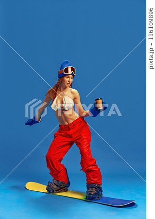 Woman combining summer and winter attire, showcasing her fit body and adventurous spirit on the slopes, standing on snowboard with coffee cup. Energy 116109896