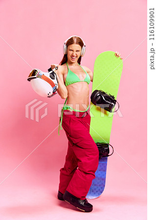 Happy and emotional young girl with tanned body, posing in winter clothes and bikini top, with snowboarding equipment against pink studio background 116109901