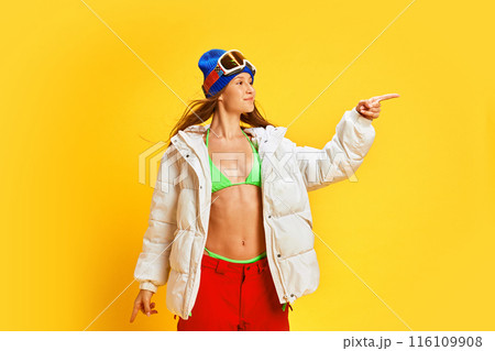 Happy woman in unique mix of winter sports attire and summer wear, choosing her travelling destination against bright yellow studio background Happy woman in unique mix of winter sports attire and summer wear, choosing her travelling destination against bright yellow studio background 116109908