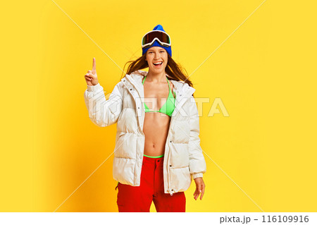 Energetic young woman in vibrant winter gear, expressing happiness and enthusiasm, against bright yellow studio background. Ready for vacation 116109916