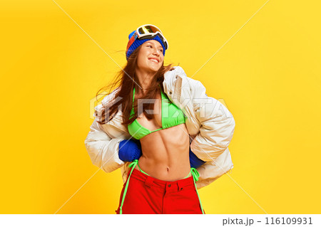 Vibrant young woman in mixed winter and summer attire, expressing excitement and fun against bright yellow studio background. Tanned body in winter season 116109931