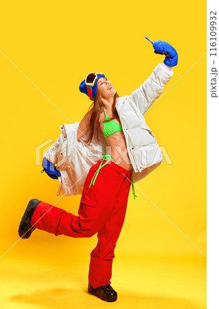 Full-length image of happy young girl in bikini top and winter clothes taking selfie n goggles against bright yellow studio background 116109932
