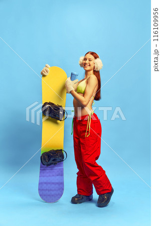 Summer Vibes in Winter Gear. Young woman in bikini top and snow pants cheerfully posing with snowboard against blue studio background 116109956