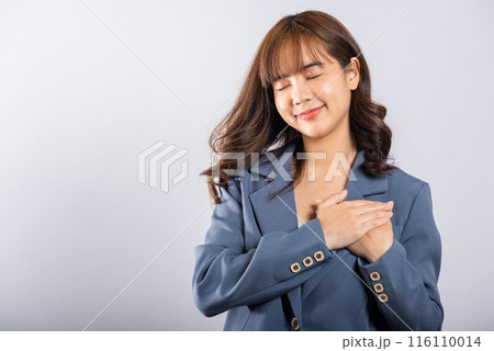 A happy business woman in casual attire holds her hands close to her chest, meditating and smiling. Studio shot isolated on a white background, portraying the concepts of belief, faith, and gratitude 116110014