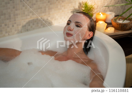 A woman releases in a bubble bath and applied a sweet face mask to her skin. She enjoys a moment of self-care and pampering in her bathroom. A woman releases in a bubble bath and applied a sweet face mask to her skin. She enjoys a moment of self-care and pampering in her bathroom. 116110881