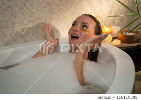 A woman is relaxing in a bubble bath with eye patches, enjoying a spa treatment. The bathroom is it with candles, creating a calming atmosphere. 116110889
