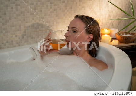 A woman enjoys a relaxing evening in a bubble bath, sipping on a glass of wine. The bath is filled with bubbles and the room is lit by candles. The woman appears to be enjoying her time and is clearly A woman enjoys a relaxing evening in a bubble bath, sipping on a glass of wine. The bath is filled with bubbles and the room is lit by candles. The woman appears to be enjoying her time and is clearly 116110907
